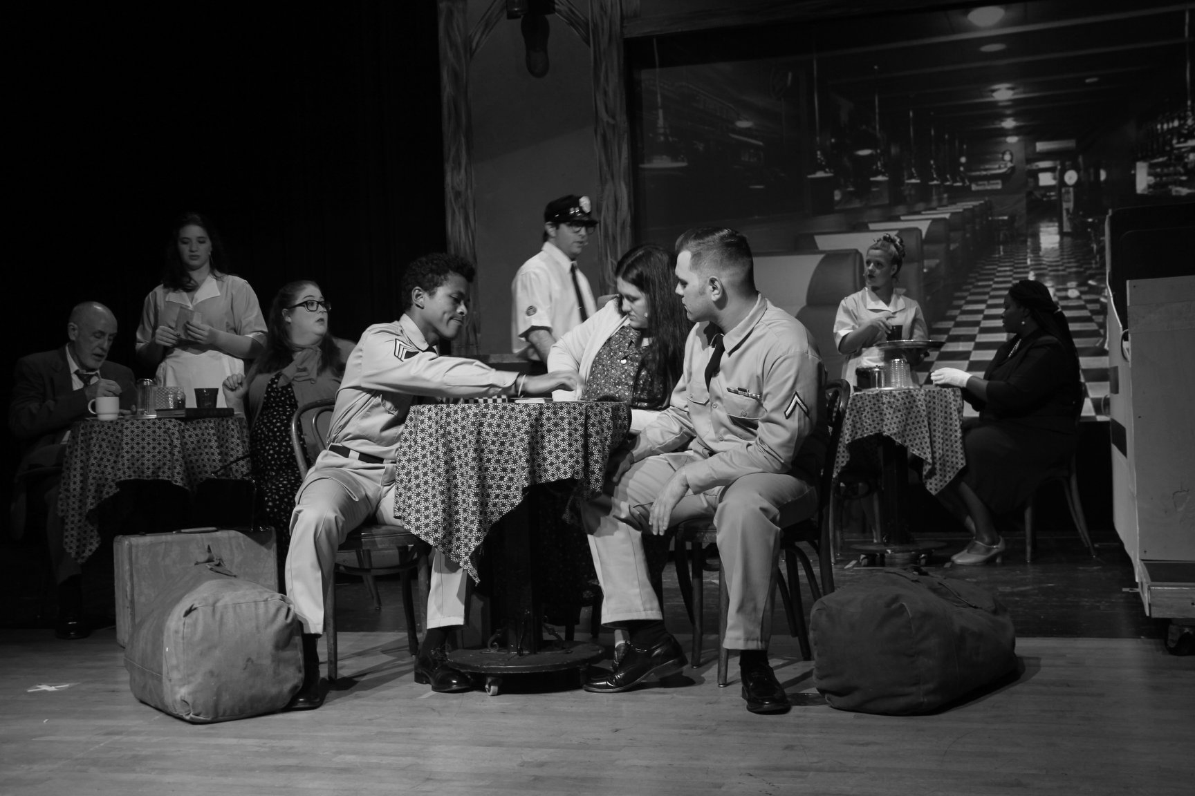 A black and white photo of a stage production. People in costumes sit at tables in a diner-like setting.