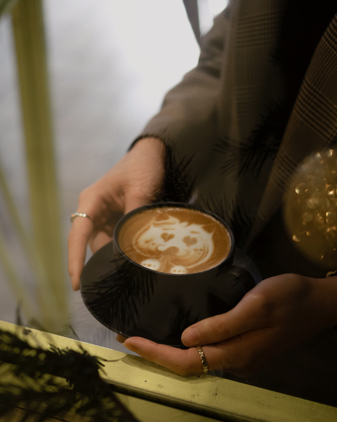 Person holding coffee cup with latte art of a cat face. Dark cup and saucer.