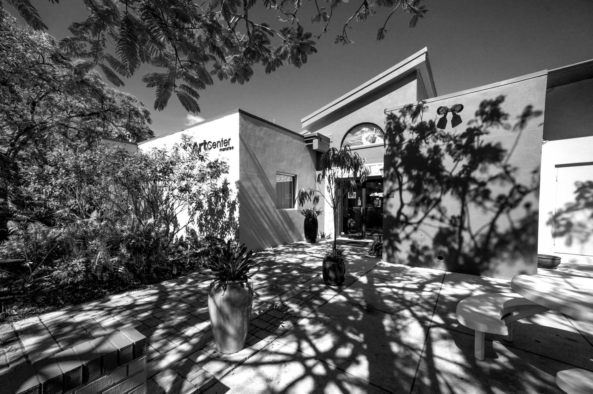 Black and white photo of a building entrance shaded by trees, with a decorative vase and outdoor seating visible.