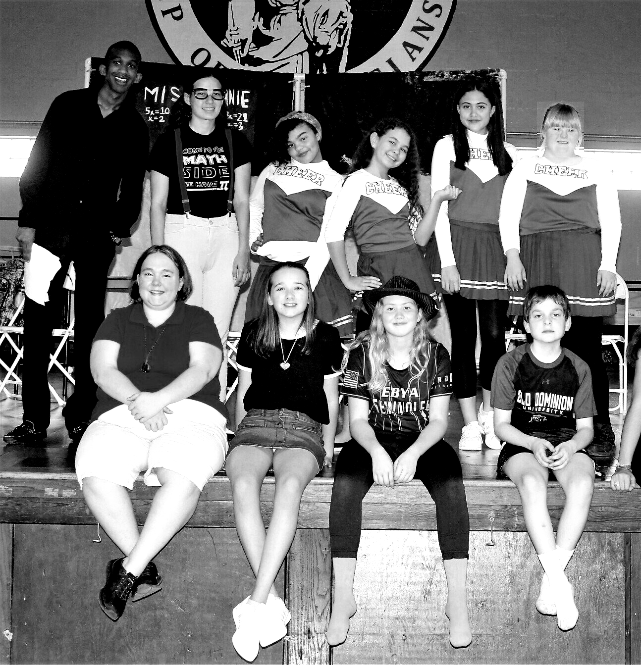 Group of people, mostly children, on a stage. Some are in cheerleading outfits, others in casual attire.