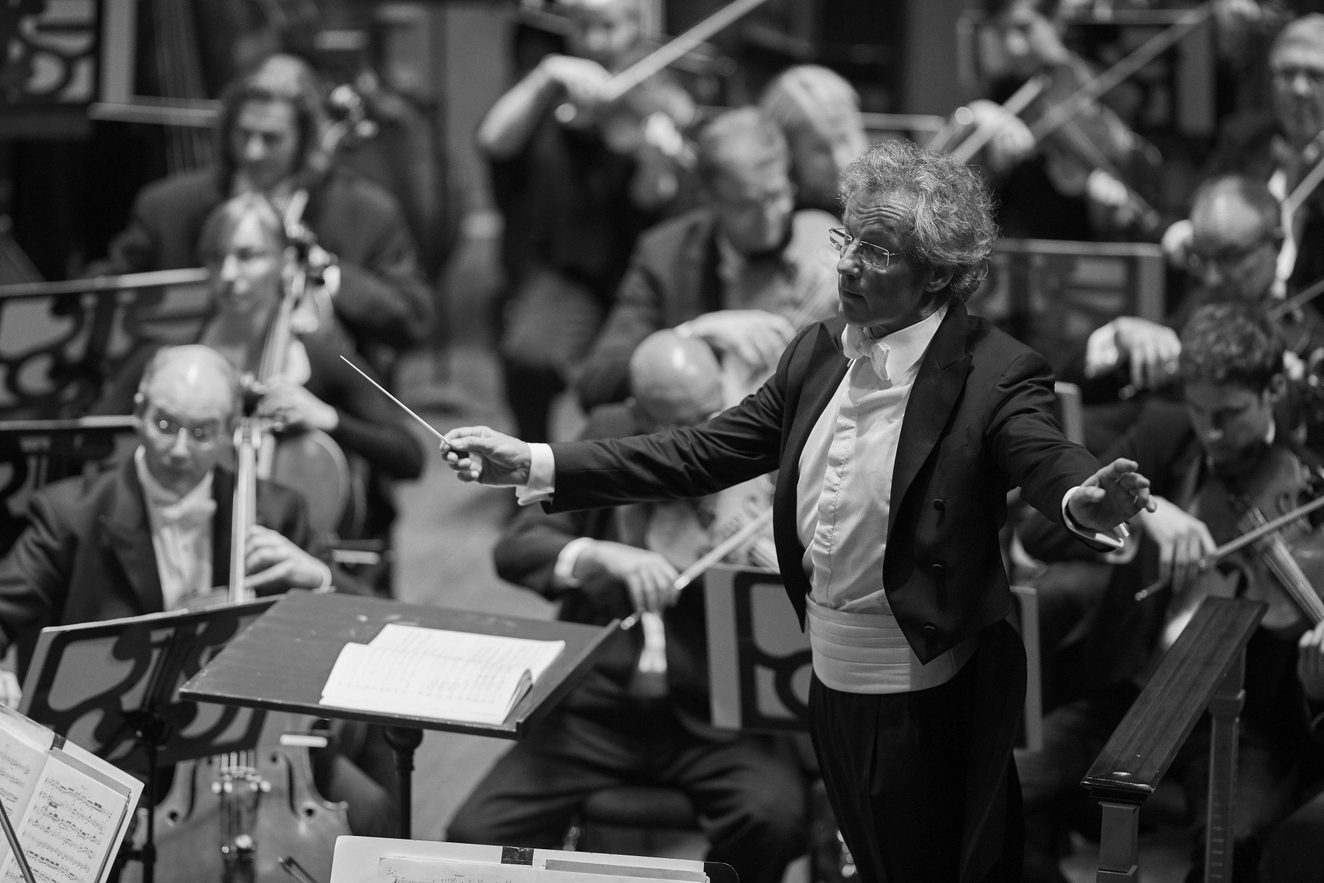 A conductor leading an orchestra, arms raised, in a concert hall. Black and white.