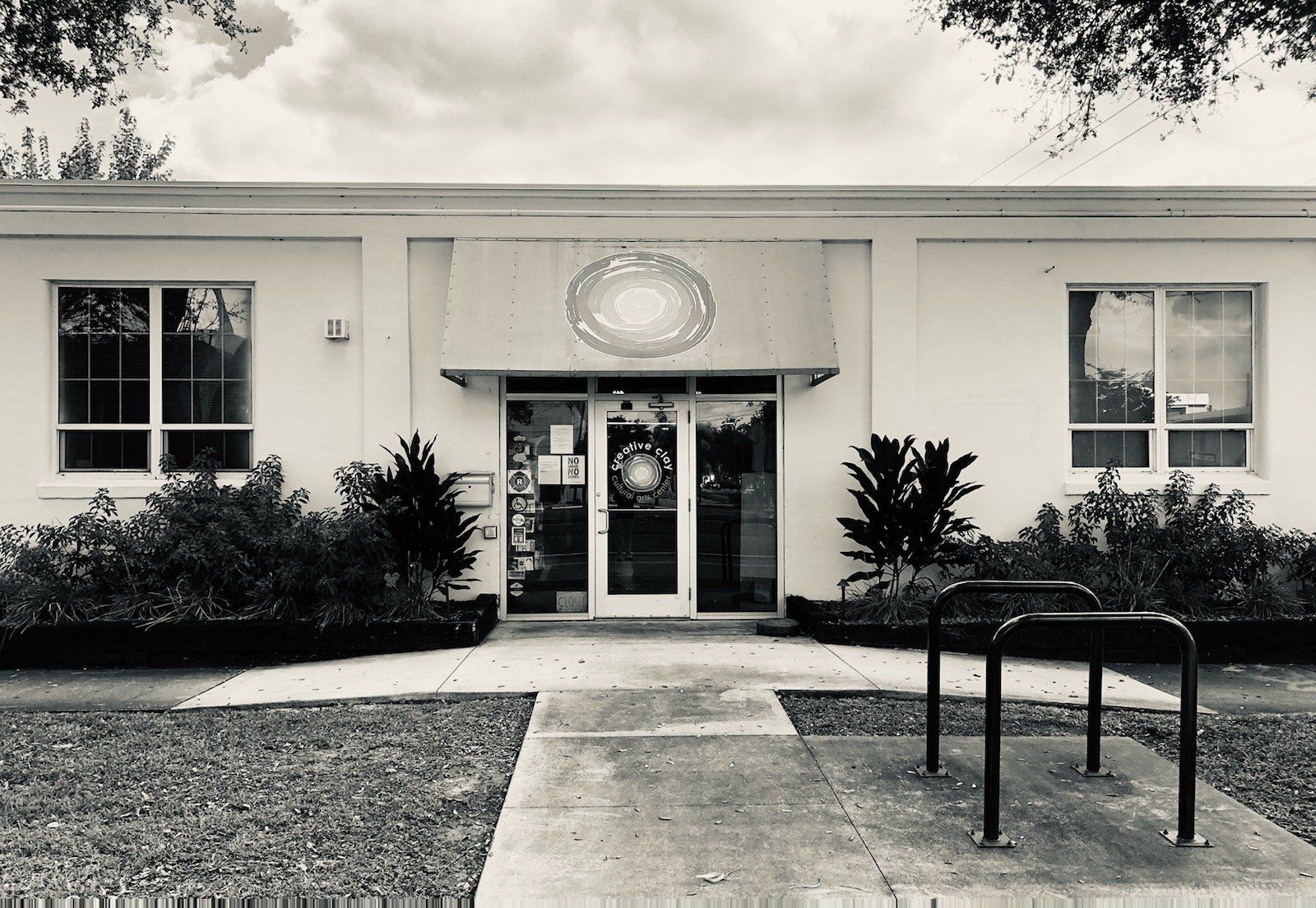 Black and white photo of a light-colored building with a front door and windows. Bushes and bike rack in front.