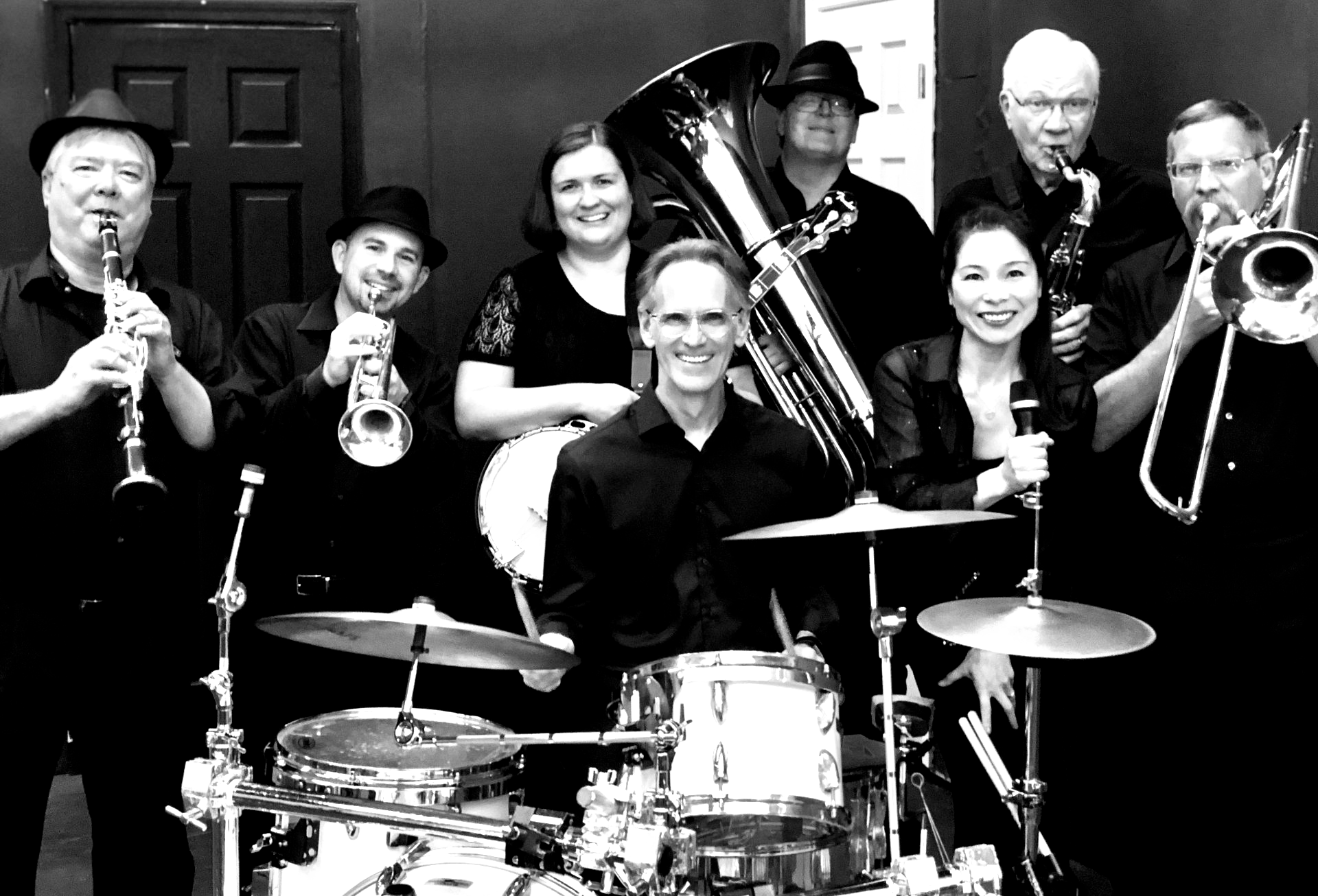 Band of nine people in black clothing pose together. Musicians: clarinet, trumpet, drums, trombone, tuba, saxophone, female vocalist.
