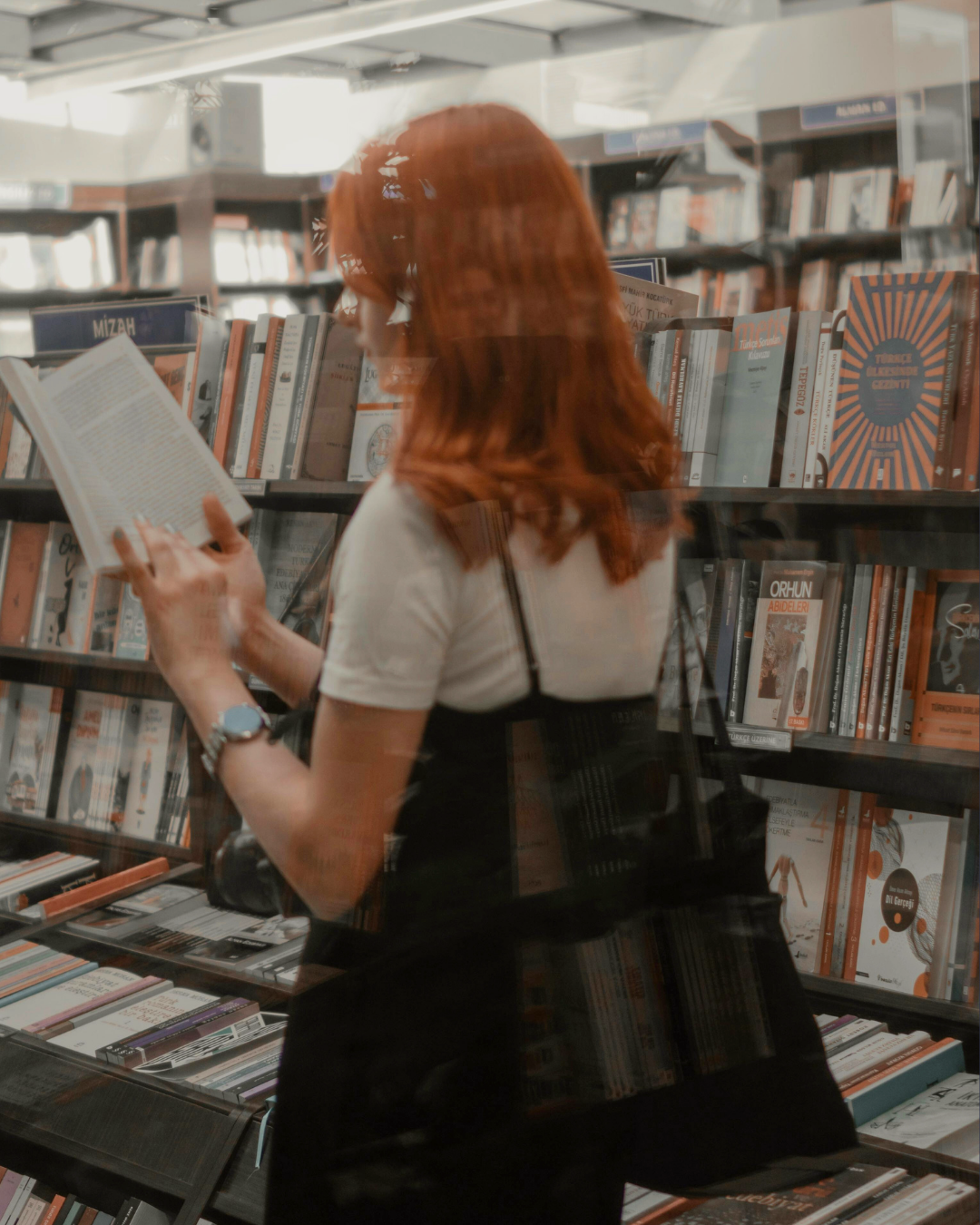 Woman with red hair in a bookstore, looking at a book. She wears a white shirt, black dress, and carries a black bag.