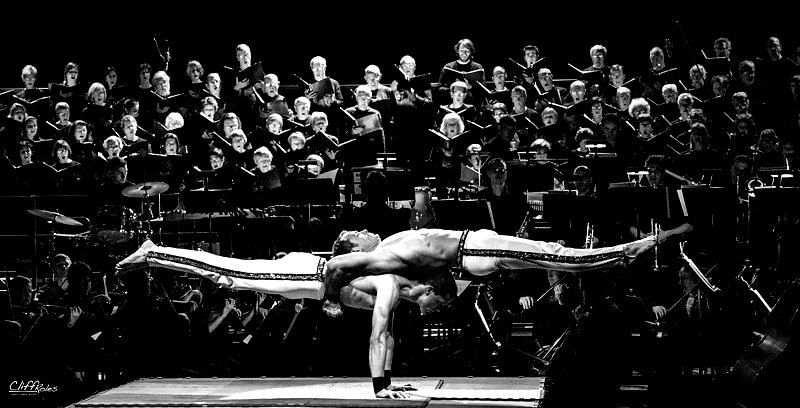Acrobats performing a balancing act on stage, with a choir and orchestra in the background.