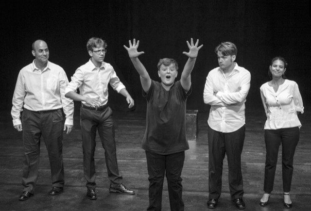 Group of five people on stage: one with arms raised, others in white shirts, dark pants.