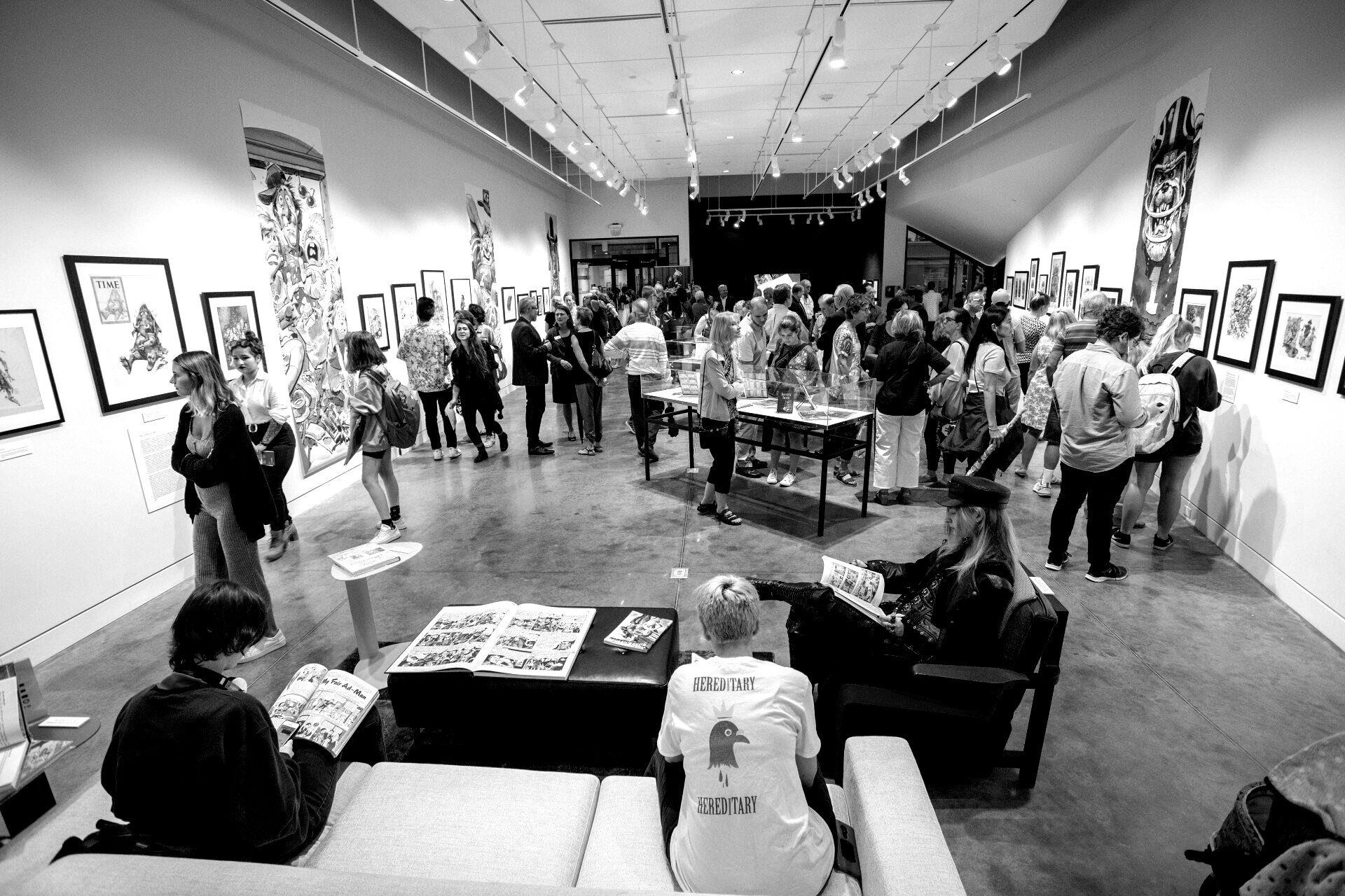 Black and white photo of crowded art gallery opening. People viewing art, talking, and sitting on couches.