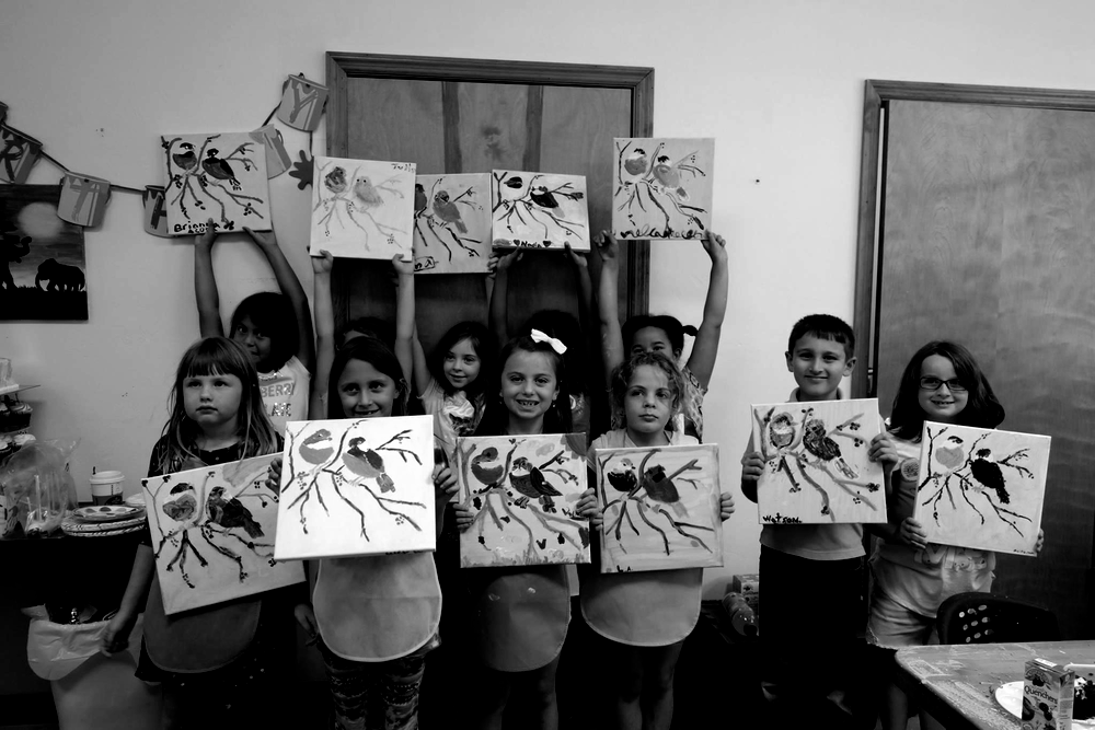 Children holding up paintings, smiling in an art classroom.