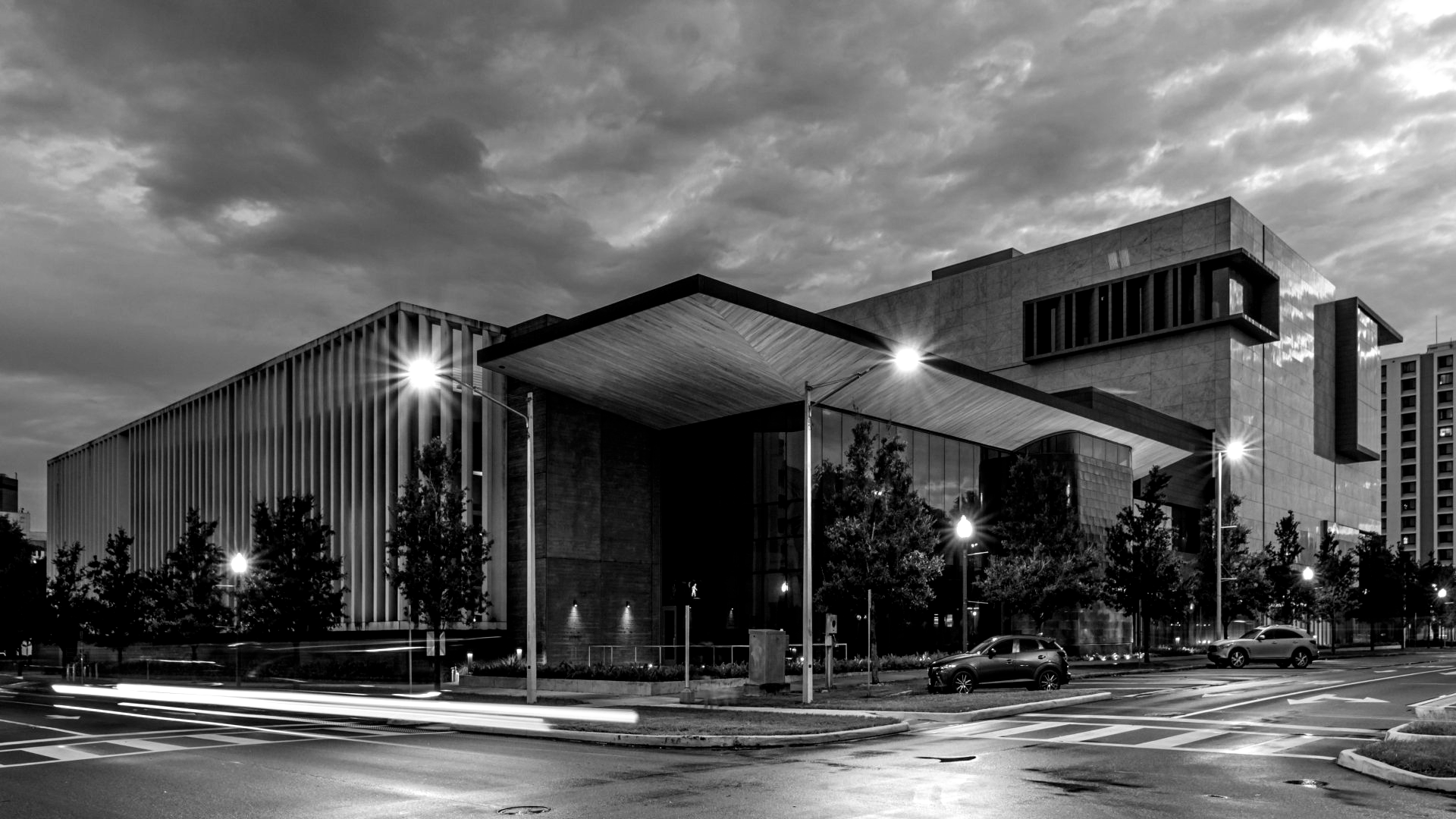 Black and white exterior shot of a large, modern building at dusk. Streetlights and cars are visible.