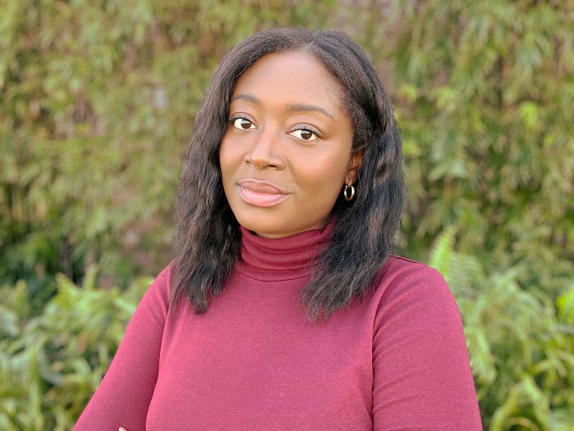 Woman with dark skin, wearing a red turtleneck, smiles slightly, outdoors against leafy green background.