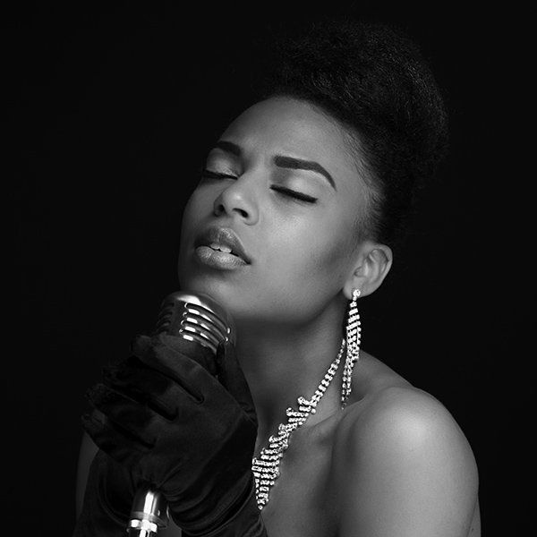 Black and white photo of a Black woman singing into a vintage microphone, eyes closed. She wears a sparkly necklace and gloves.