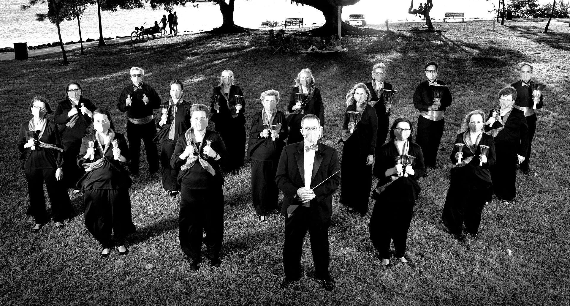 Black and white photo of choir, facing camera in park. Most hold objects, some dark clothing.