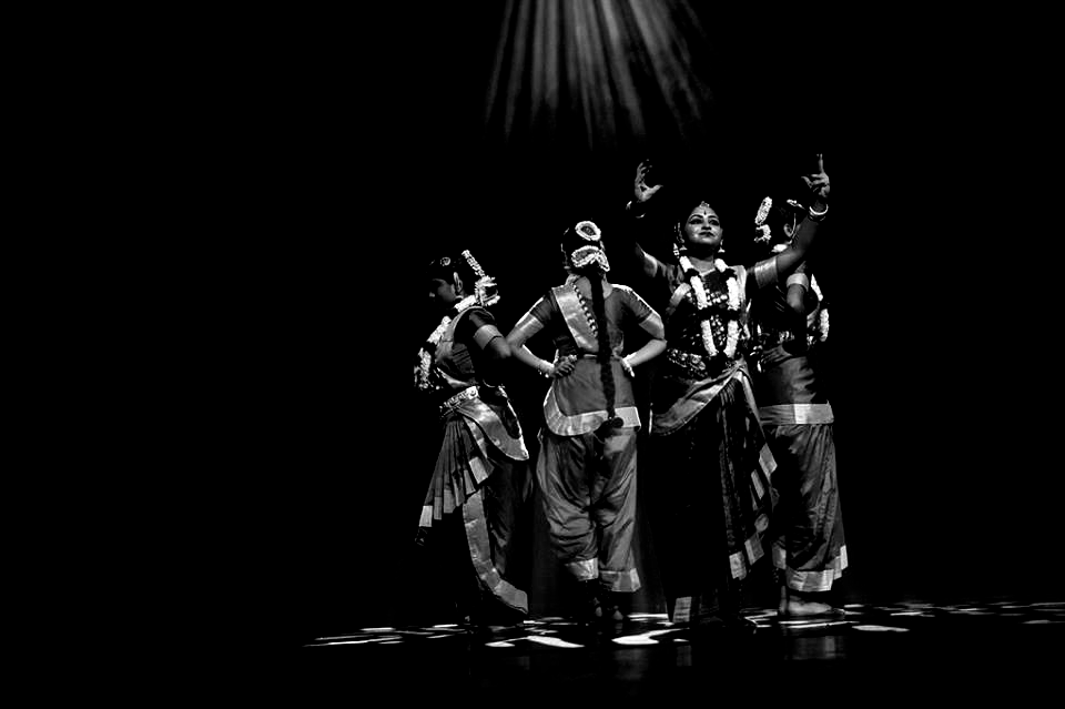 Four dancers on stage, performing with raised arms and traditional clothing, lit by a spotlight.