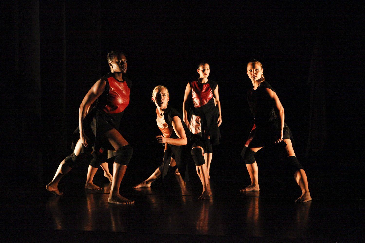 Dancers in dark costumes with red accents perform on a stage; low lighting.