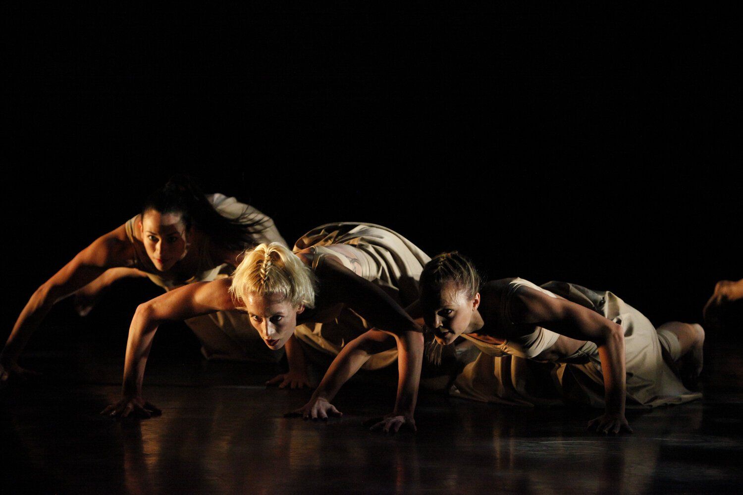 Dancers crawling low, illuminated by a spotlight, in a dark space. They wear light-colored attire, arms extended.