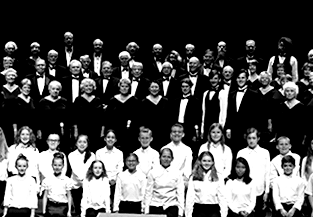 Choir performing on stage in formal attire, including adults and children, under stage lights.