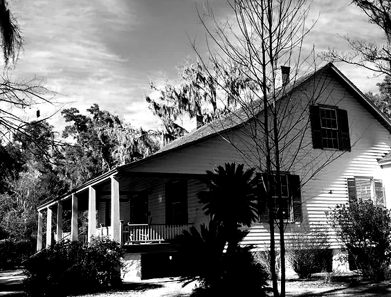 Black and white photo of a house with a porch, surrounded by trees.