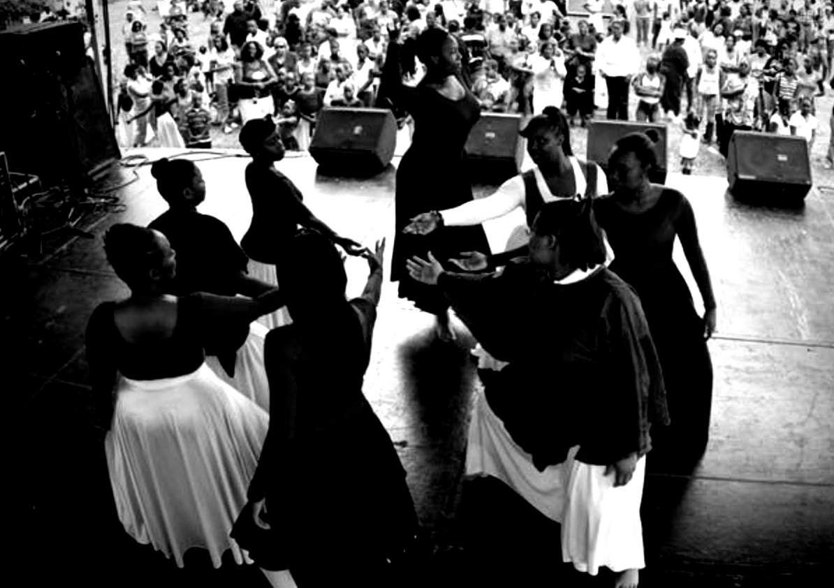 Black and white photo of a dance performance outdoors on a stage with an audience. Dancers in dark and white clothing.