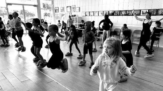 Kids in a dance class, leaping and moving on special platform shoes in a brightly lit room.