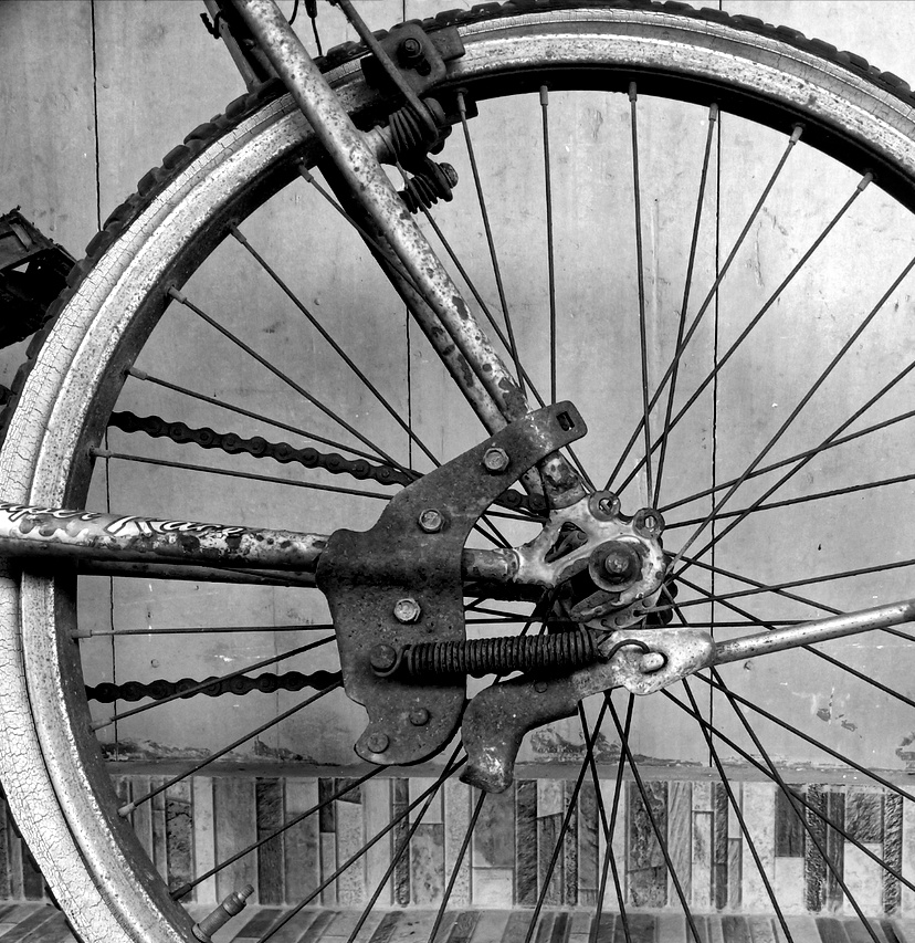 Black and white close-up of an antique bicycle wheel, showing spokes, chain, and rusted metal components.