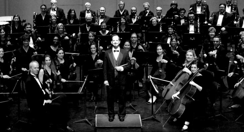 A conductor leads a large orchestra, all dressed in black and white attire, in a concert hall setting.