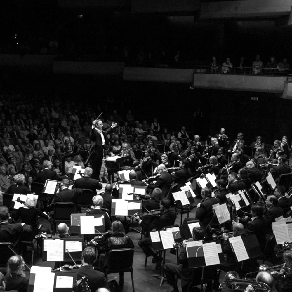A conductor leads an orchestra in a concert hall, with a large audience watching. Black and white.