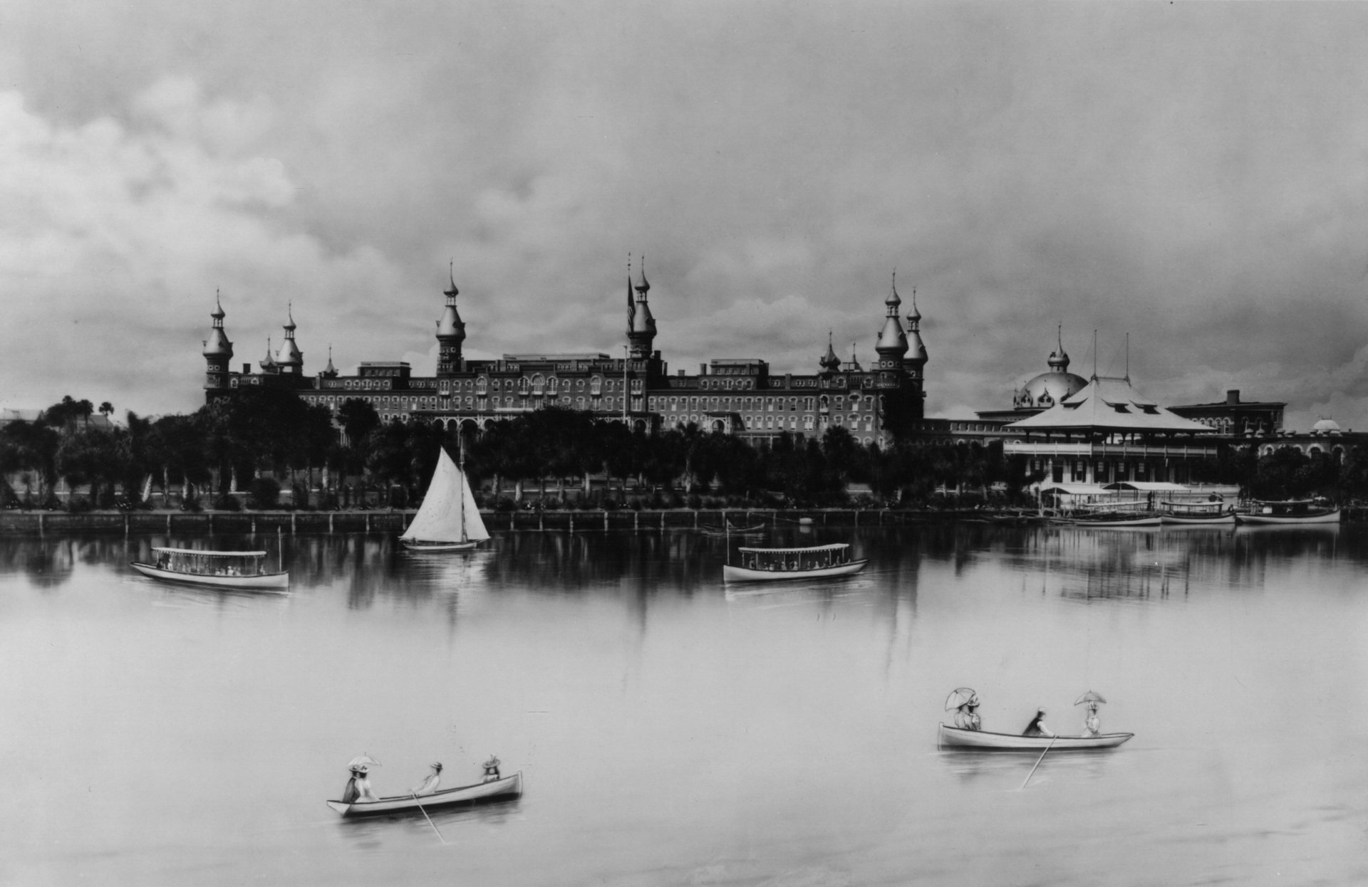 View of a large Moorish-style building on a lake with boats. Cloudy sky.