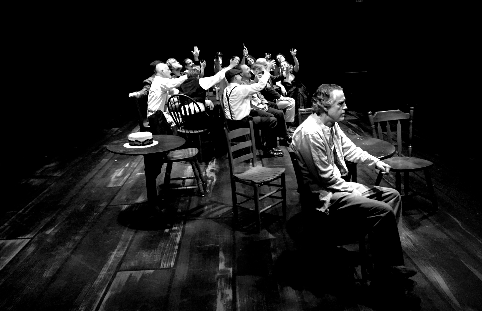 Black and white stage scene; man sits, others cheer with raised hands, tables and chairs on wooden floor.