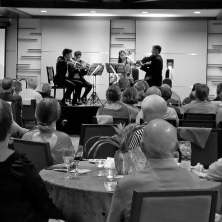 String quartet performing for an audience seated at round tables in a room. Black and white photo.