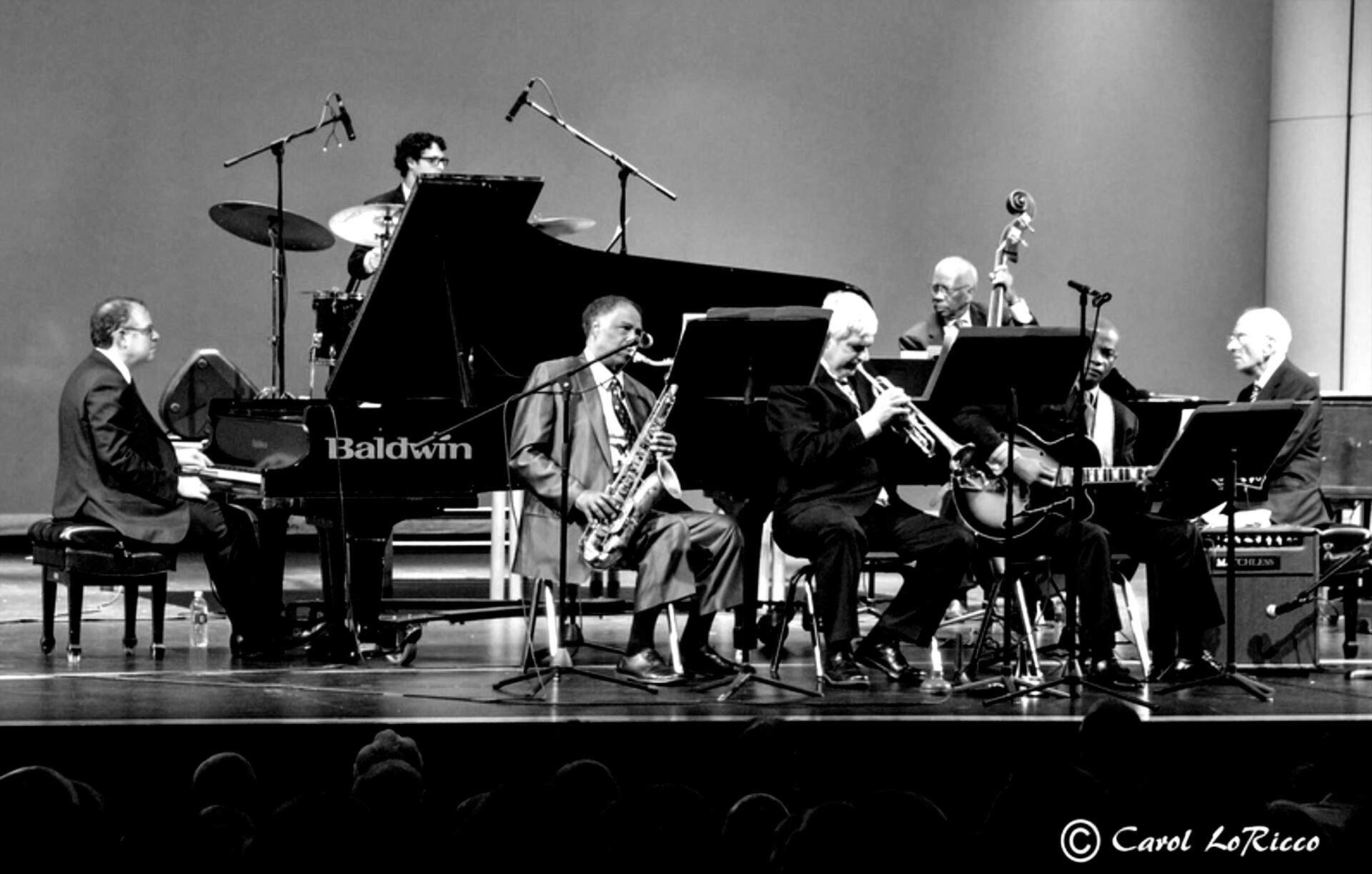 Jazz band performing on stage; black and white. Musicians include pianist, drummer, saxophonist, trumpet player, bassist, and guitarist.