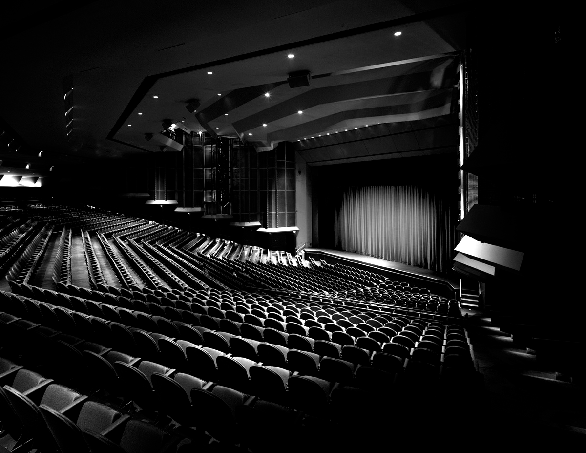 Black and white photo of an empty theater auditorium. Rows of seats face a stage with a curtain.