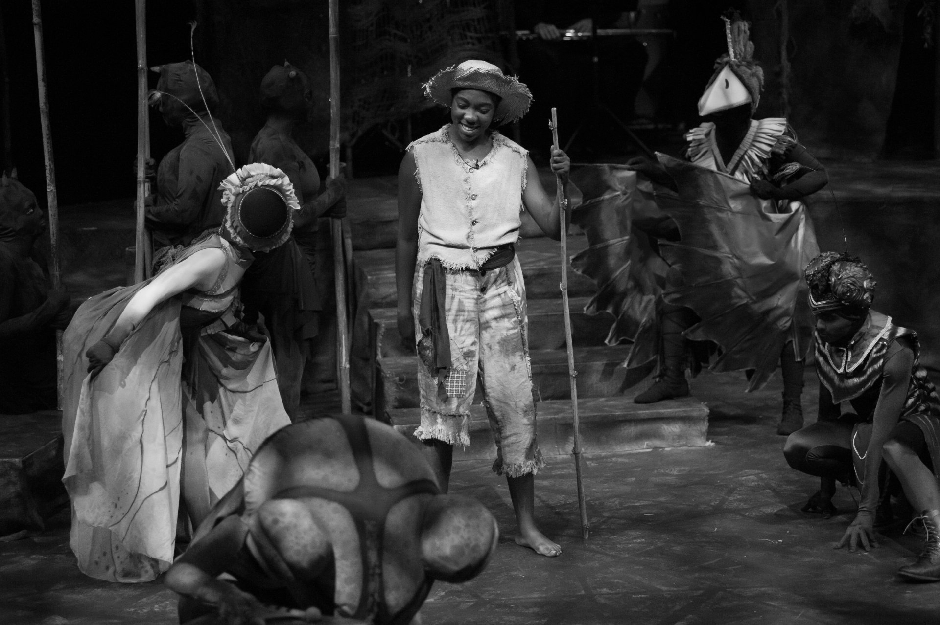 A black and white stage performance: a man smiles, others in costume surround him.