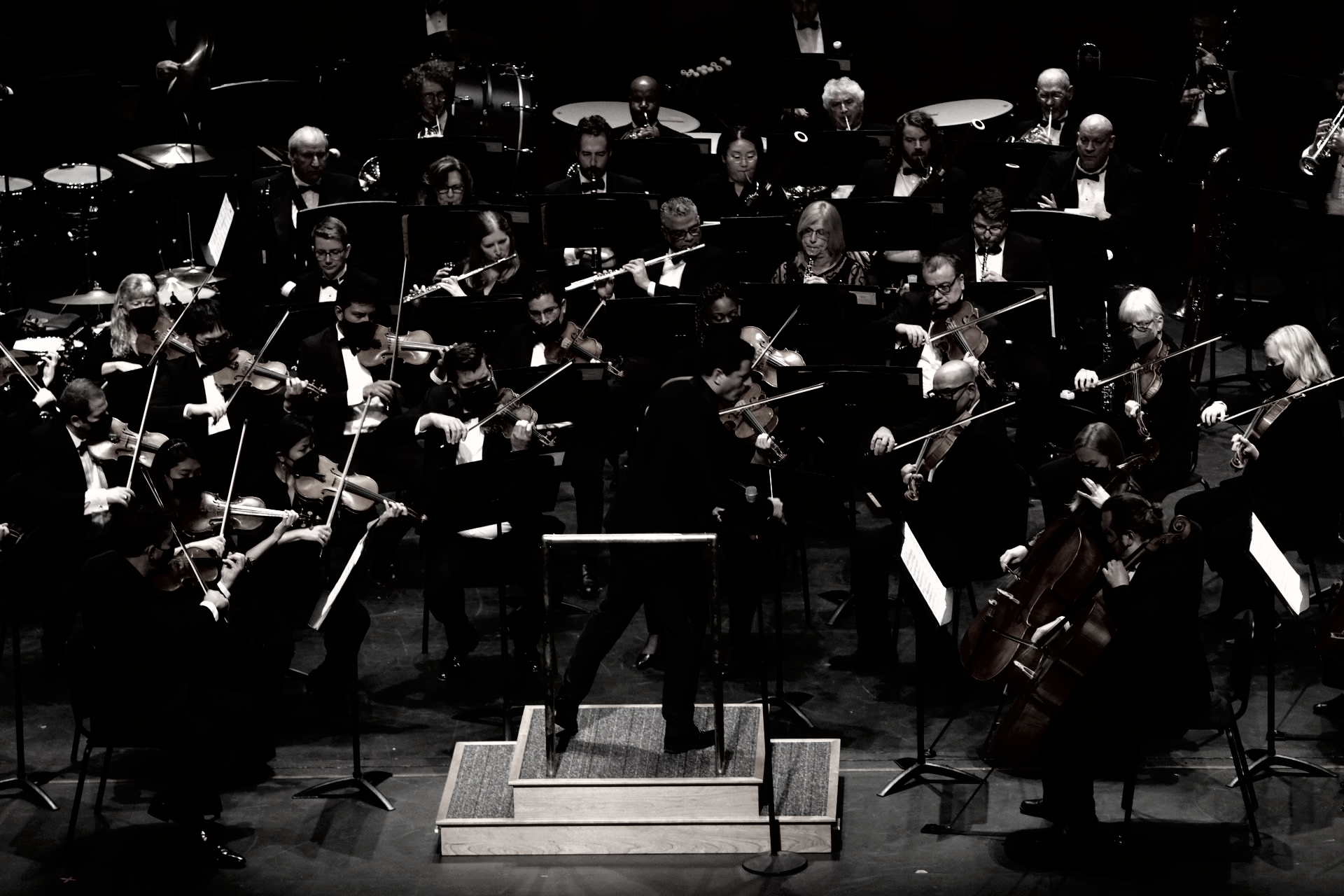Orchestra performing on stage; conductor directing musicians. Black and white.