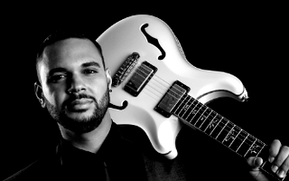 Man with a beard holding a white electric guitar against a black background.