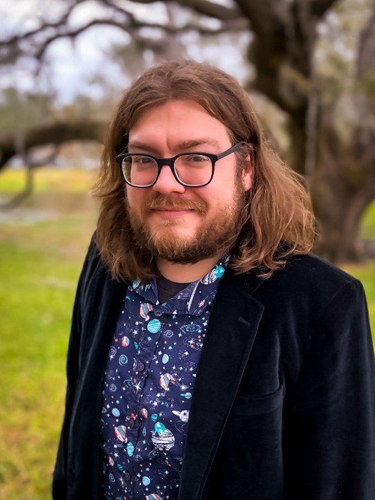 Man with long brown hair, glasses, and beard smiles outside, wearing a galaxy print shirt and dark blazer.