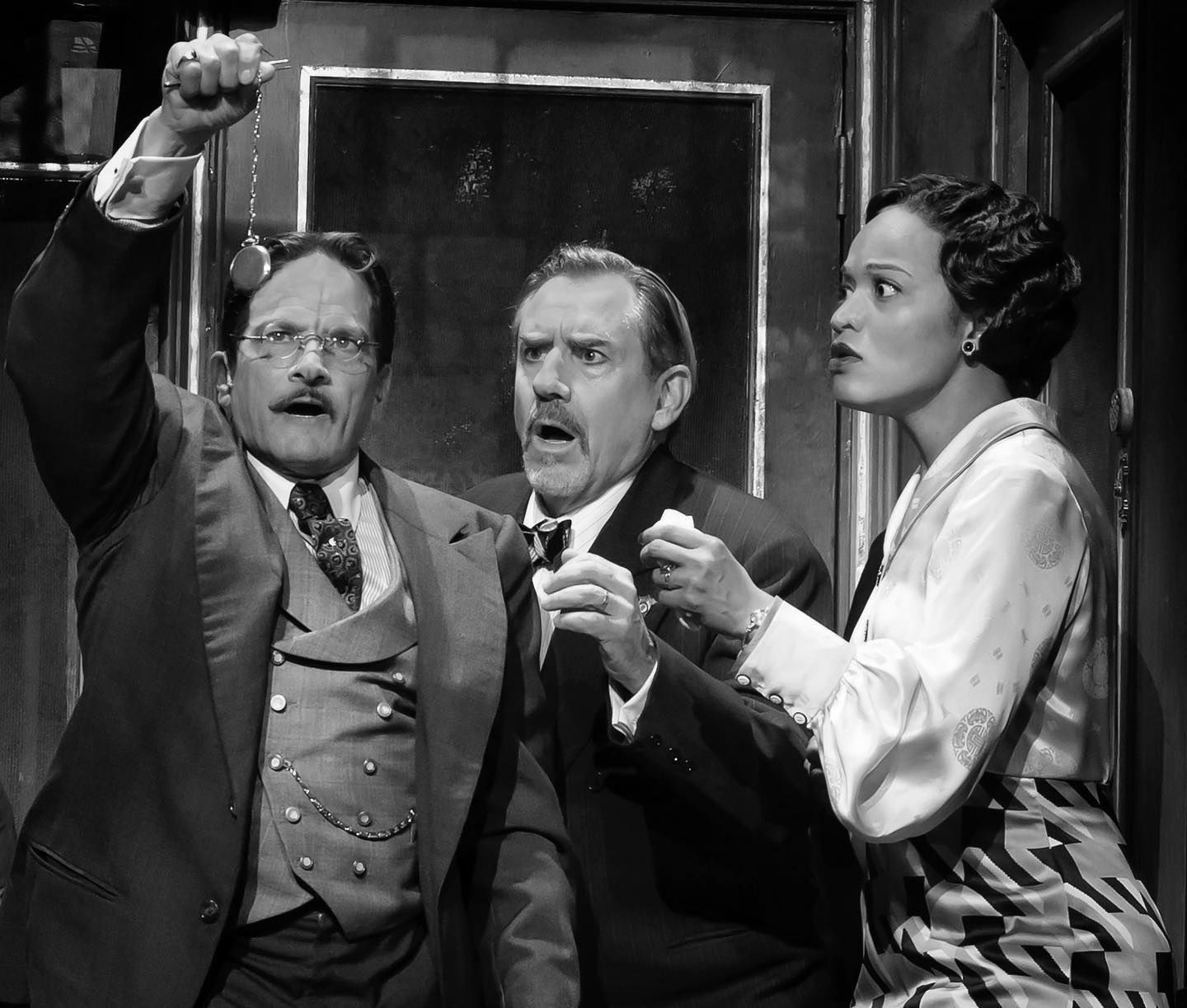 Three actors in period attire react with shock and concern. Man raises hand, another clenches fist; woman looks aghast. Dark interior.