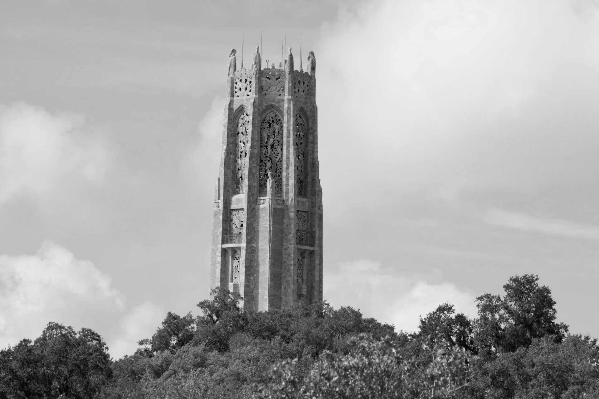 Gothic-style bell tower atop a treed hill, against a cloudy sky. Black and white.