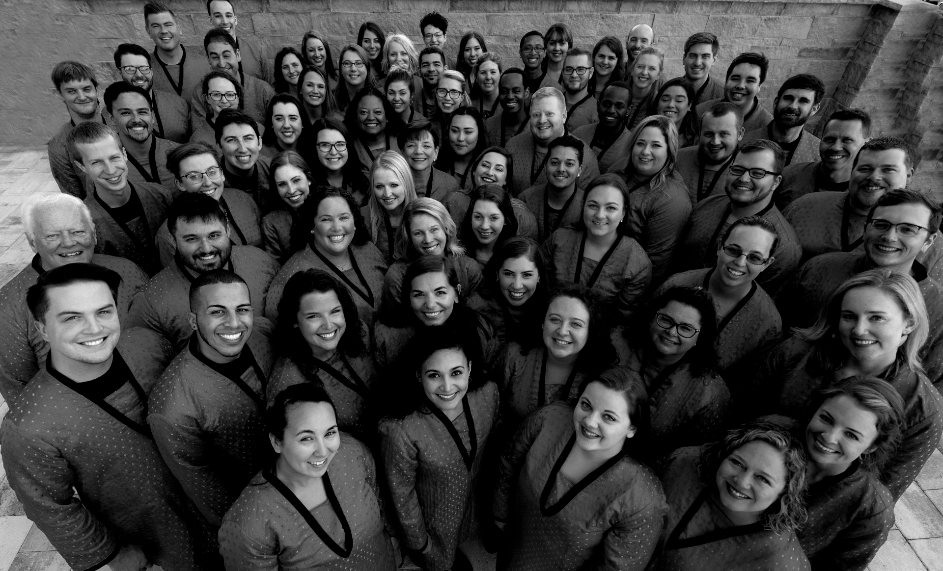 Large group of diverse people, smiling, gathered together outdoors, possibly a choir.
