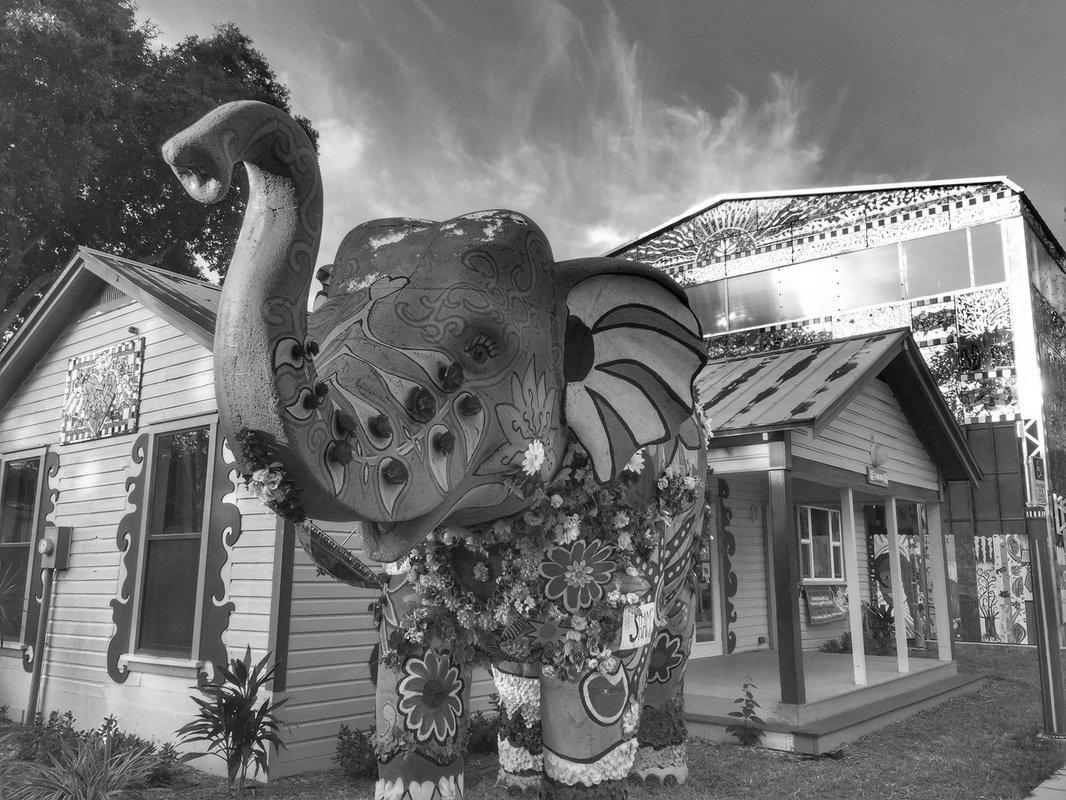 Black and white photo of a large, painted elephant statue in front of a colorful house with a porch.