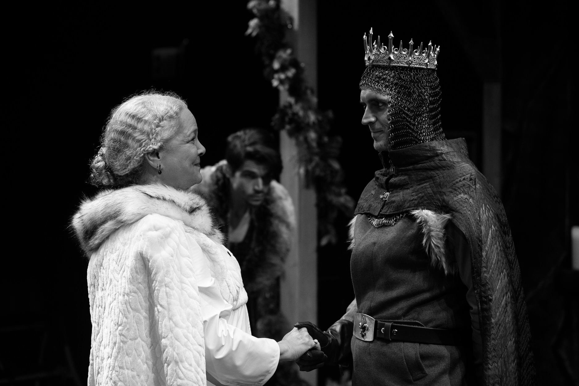 Black and white photo of a queen and king in costume, shaking hands. A man watches in the background.