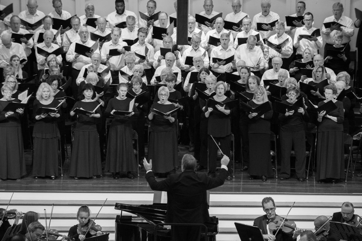 Black and white photo of a choir and orchestra performing on a stage, led by a conductor.