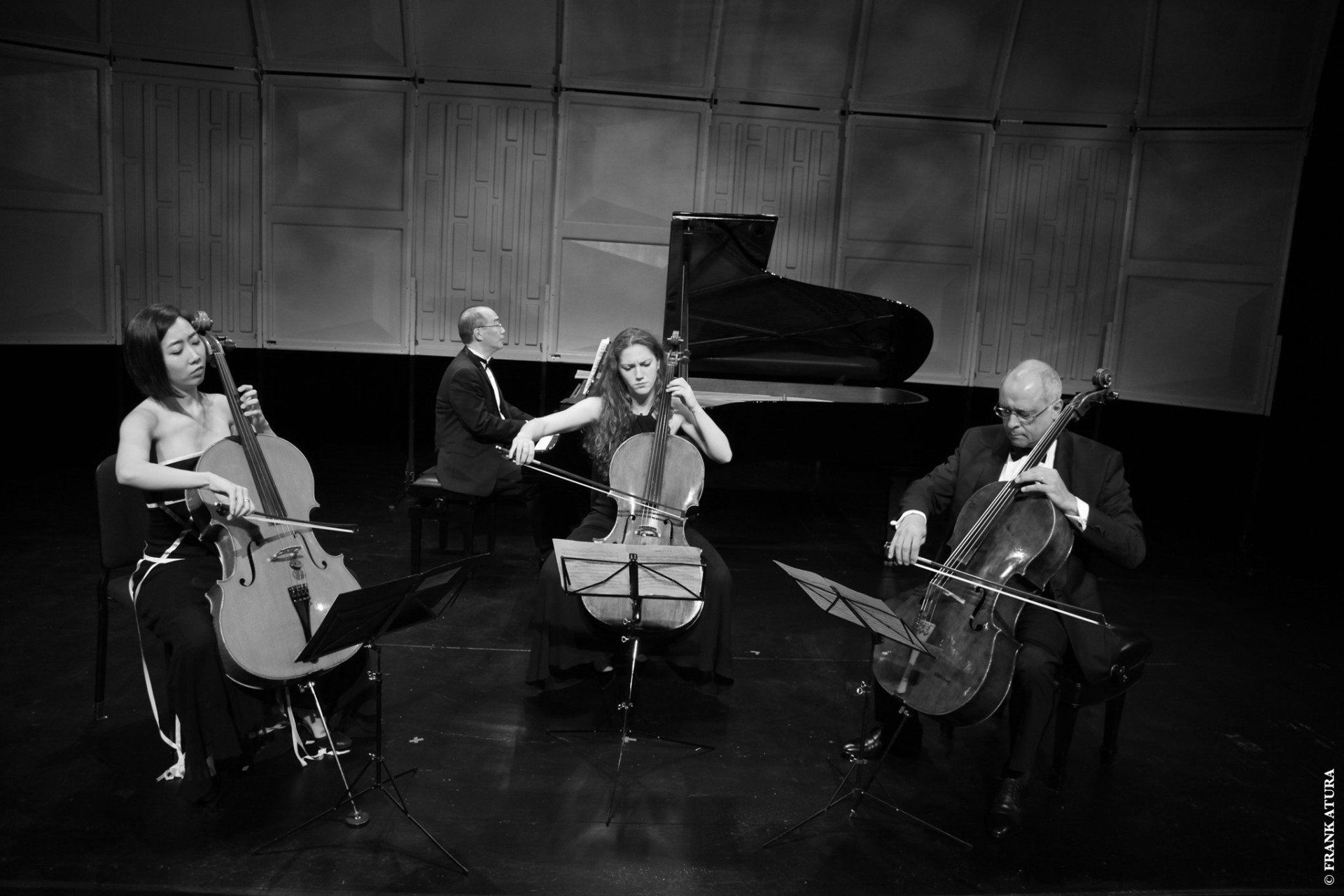 A cello quartet and a pianist perform on a stage in black and white.