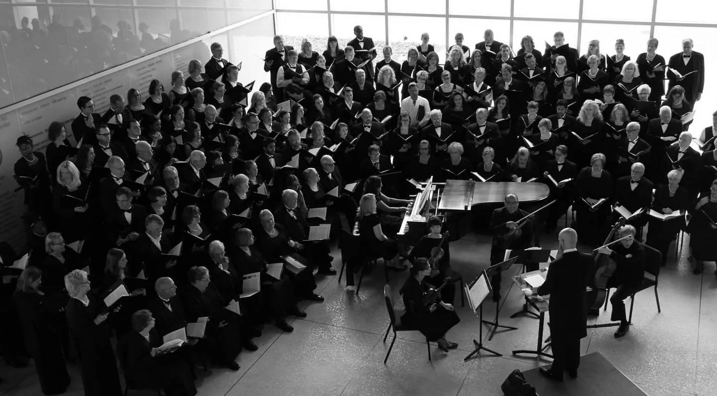 Large choir and orchestra performing in a bright hall, conducting from the front. Black and white.