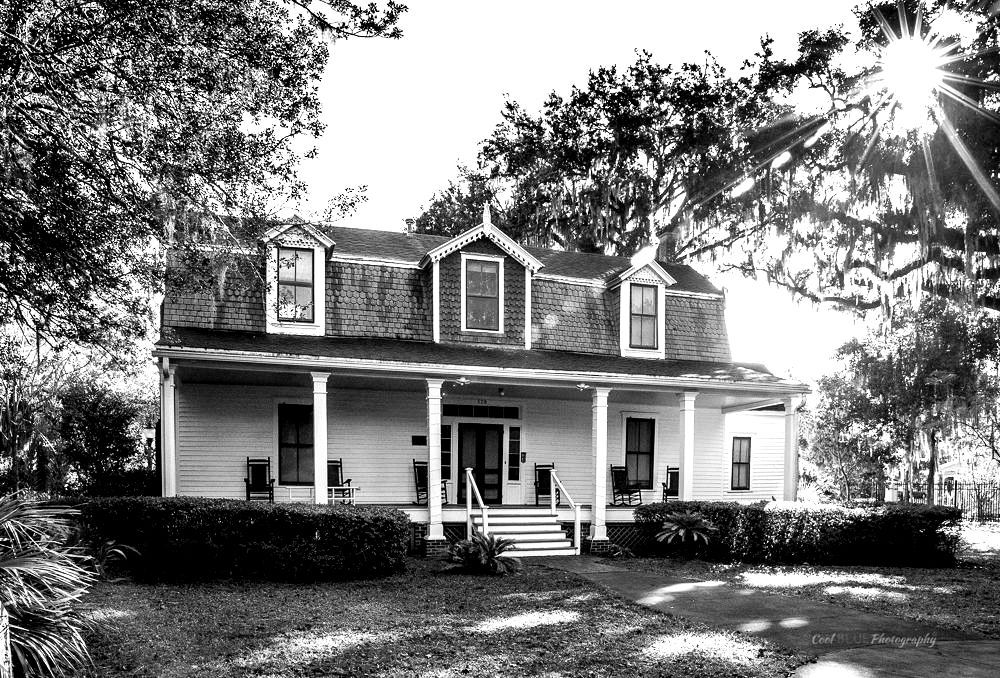 Two-story white house with a large porch, dormers, and trees in the background.