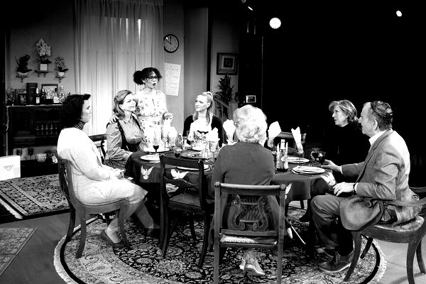 Black and white photo: a group of people seated around a dining table in a domestic setting, interacting and talking.