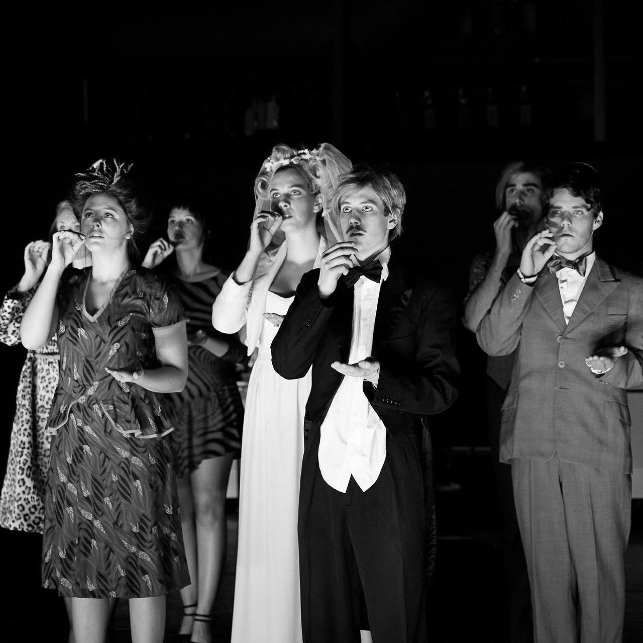 Group of people in formal wear, blowing on glass tubes. Dark room, focused light.