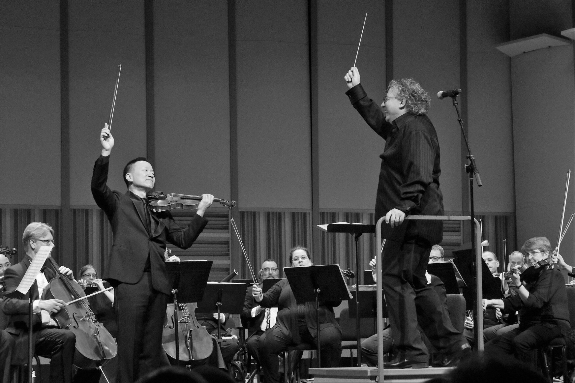 Two conductors lead an orchestra on a stage. One plays violin, raising bow. The other conducts from a podium.