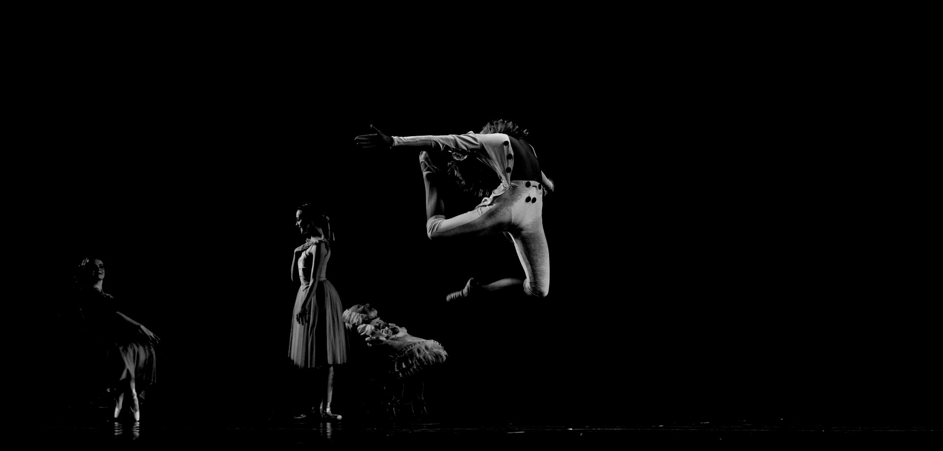 A dancer leaps mid-air in a black and white performance. Two other dancers are visible in the background.