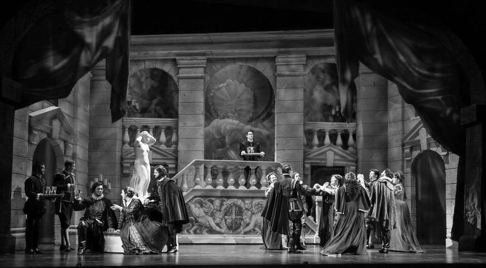 Black and white stage set; actors in period costumes, performing on a balcony with statues and columns.