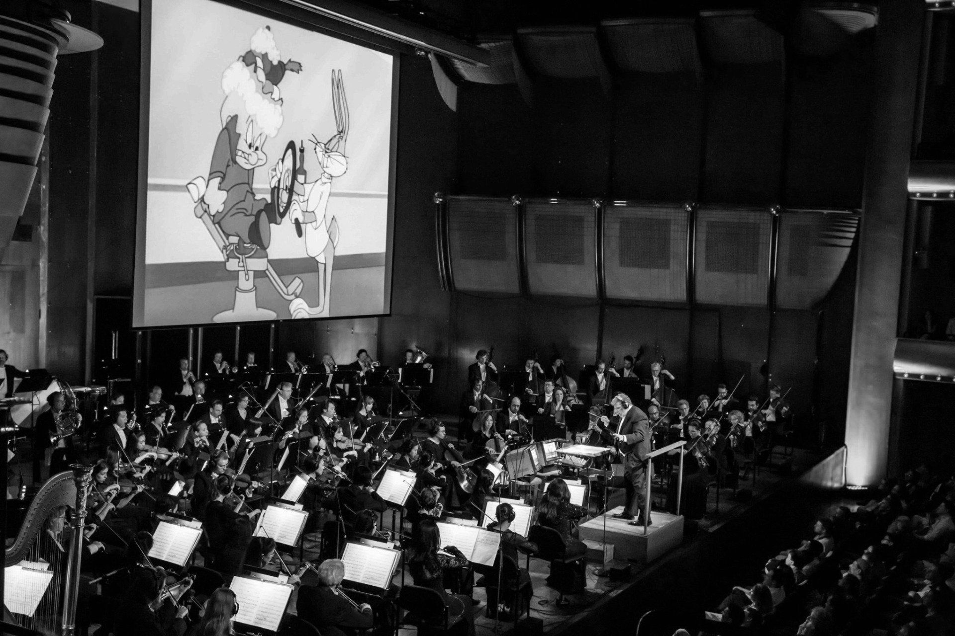 Black and white photo of orchestra performing with cartoon projected on a screen above.
